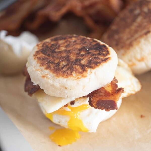 sourdough breakfast sandwich made with English muffins, crispy bacon, sharp cheddar cheese, and an over easy egg with the yolk that has broken and running down the sandwich. The sandwich sits on parchment paper with more ingredients in the background