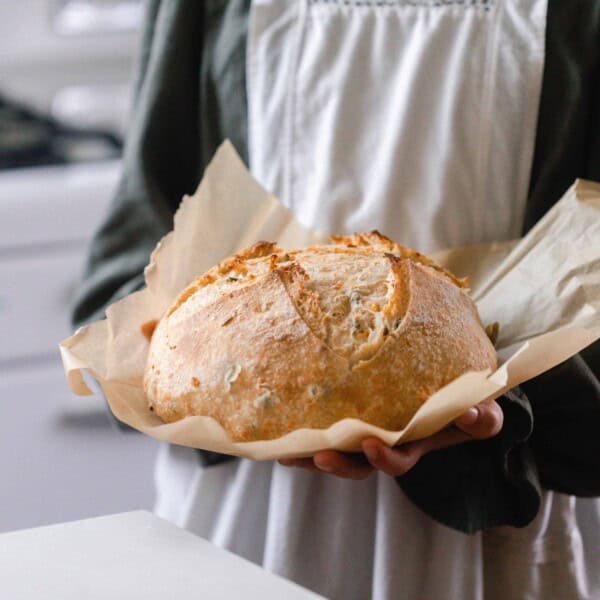 girl in a green dress with a gray apron holding a loaf of cheddar jalapeño sourdough bread on parchment paper