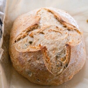 overhead photo of a crusty artisan loaf of cheddar jalapeño sourdough bread on parchment paper with a black and white stripped towel to the left