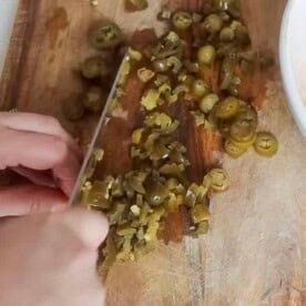 hand chopping pickled jalapeños on a wood cutting board with a bowl of shredded cheese to the left.