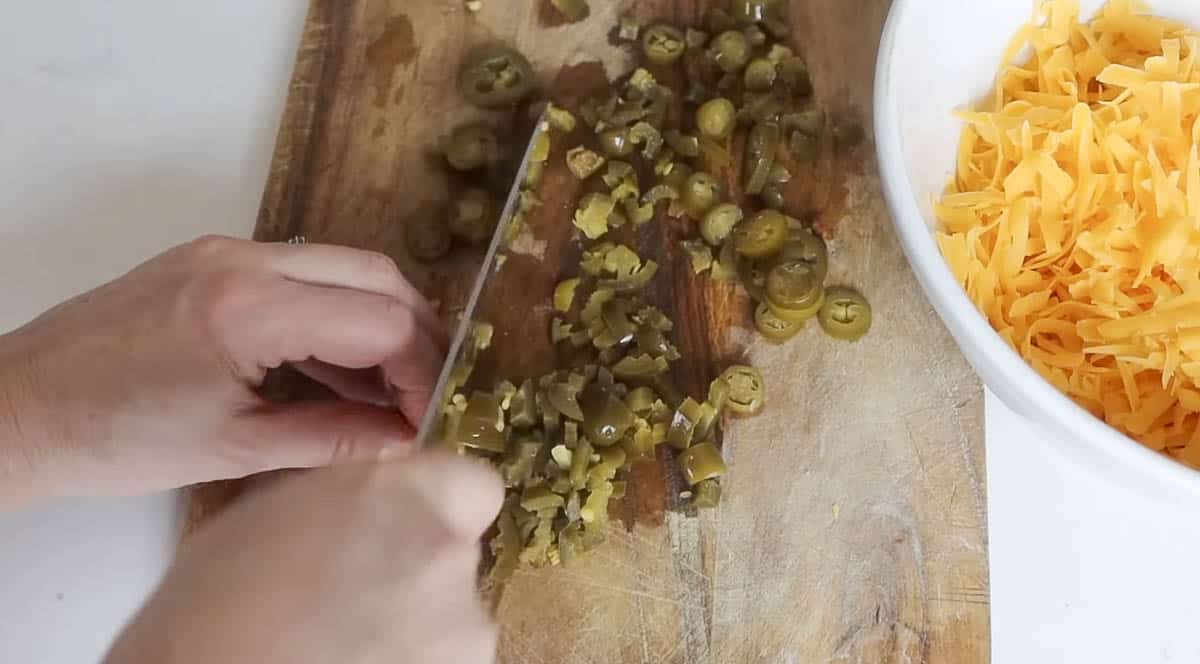 hand chopping pickled jalapeños on a wood cutting board with a bowl of shredded cheese to the left.