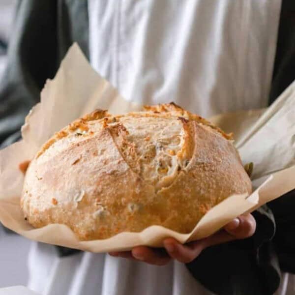 girl in a green dress with a gray apron holding a loaf of cheddar jalapeño sourdough bread on parchment paper