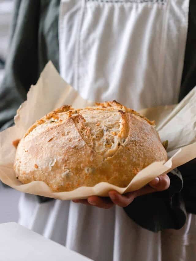 girl in a green dress with a gray apron holding a loaf of cheddar jalapeño sourdough bread on parchment paper