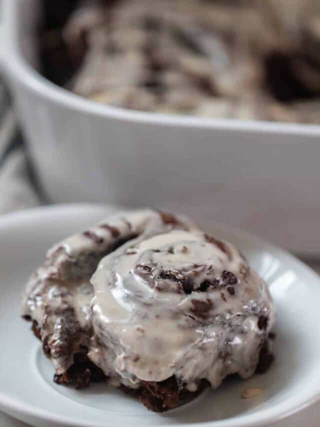 double chocolate sourdough roll with vanilla icing on a white plate with a white baking dish in the background with more rolls