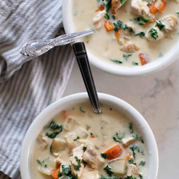 Two bowls of chicken kale soup with a silver spoon in each on a white countertop with a blue and white striped tea towel on the side