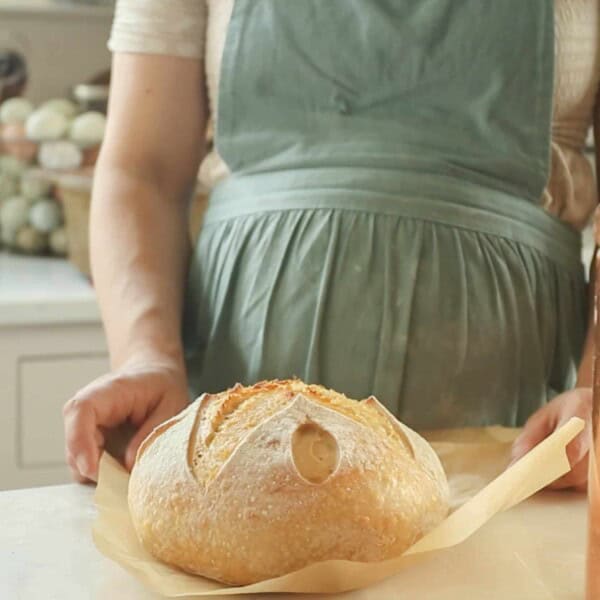 a sourdough boule on parchment paper on a white countertop with a woman in a green apron. With a copper canister to the left