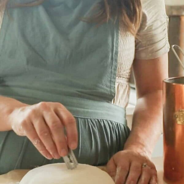 a woman wearing a green apron scoring a sourdough boule on a countertop with parchment paper