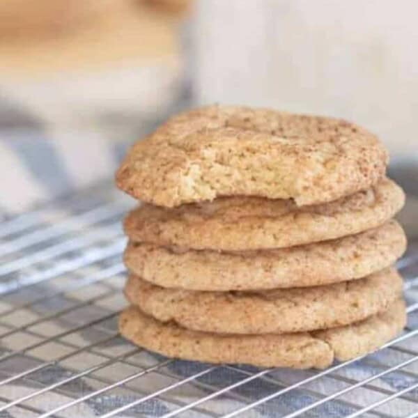 five sourdough snickerdoodle cookies stacked up on a wire rack over a white and blue stripped towel with a glass of milk behind the stack of cookies