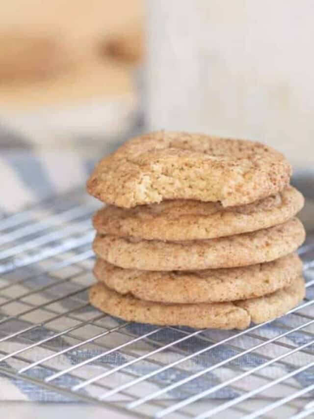 five sourdough snickerdoodle cookies stacked up on a wire rack over a white and blue stripped towel with a glass of milk behind the stack of cookies