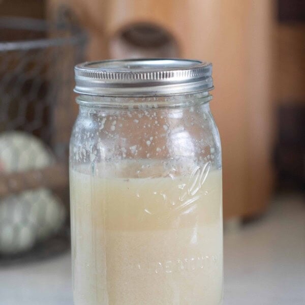 A glass jar of potato flake sourdough starter with a grain mill and eggs in the background on a white counter top