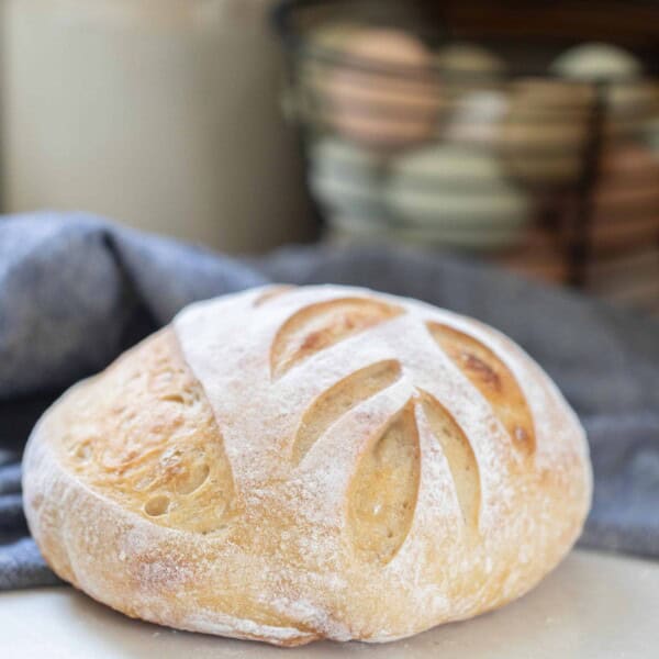 loaf of same day sourdough bread with a leaf pattern on a white counter with a blue towel and a basket of eggs in the background