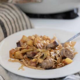 A white bowl filled with sausage and sauerkraut with a silver fork in it and a white and beige tea towel in the background