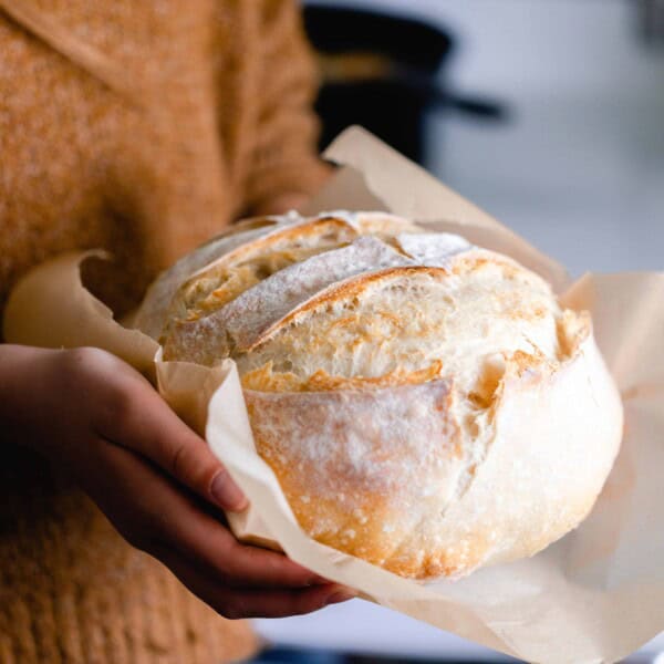 girl wearing a burn orange sweater holding a loaf of sourdough bread with parchment paper on the bottom of the loaf