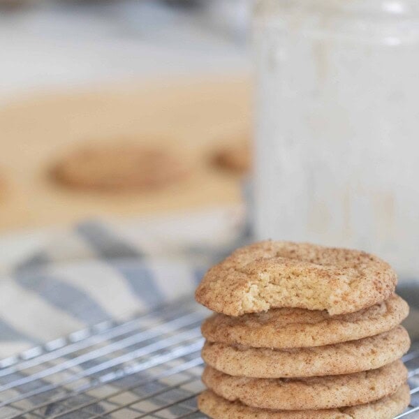 five sourdough snickerdoodle cookies stacked up on a wire rack over a white and blue stripped towel with a glass of milk behind the stack of cookies