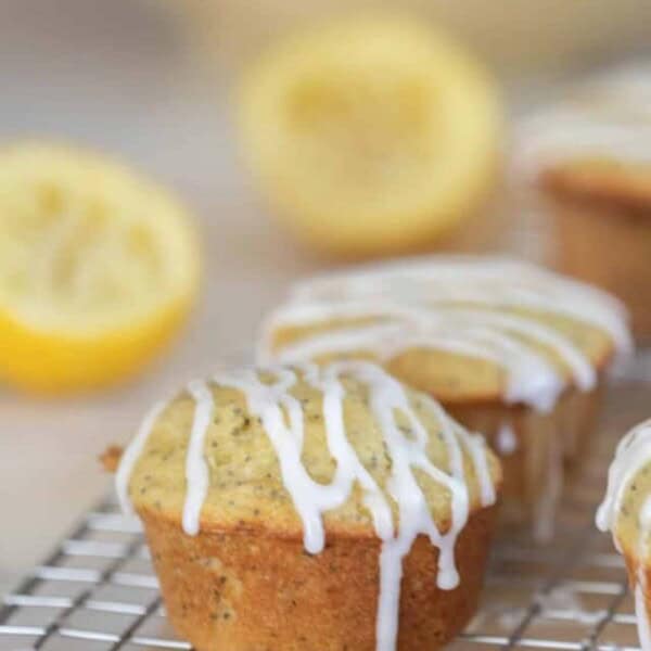 ten lemon poppy seed sourdough muffins with a lemon glaze drizzled over top on a wire rack with two lemons and a large bowling the background