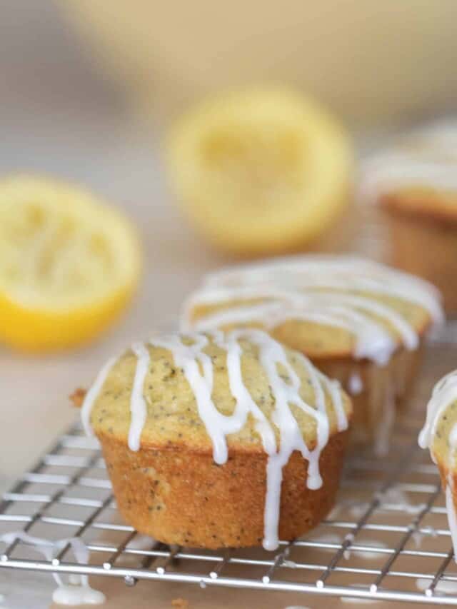 ten lemon poppy seed sourdough muffins with a lemon glaze drizzled over top on a wire rack with two lemons and a large bowling the background