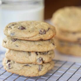 four sourdough chocolate chip cookies stack up on top of each other on a wire rack with a glass of milk and more cookies in the background