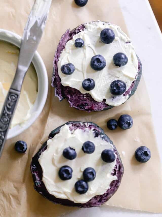 overhead photo of a sourdough blueberry bagel sliced in half and topped with cream cheese topped with blueberries. The bagels are on parchment paper with a cream cheese in a white container and topped with a knife