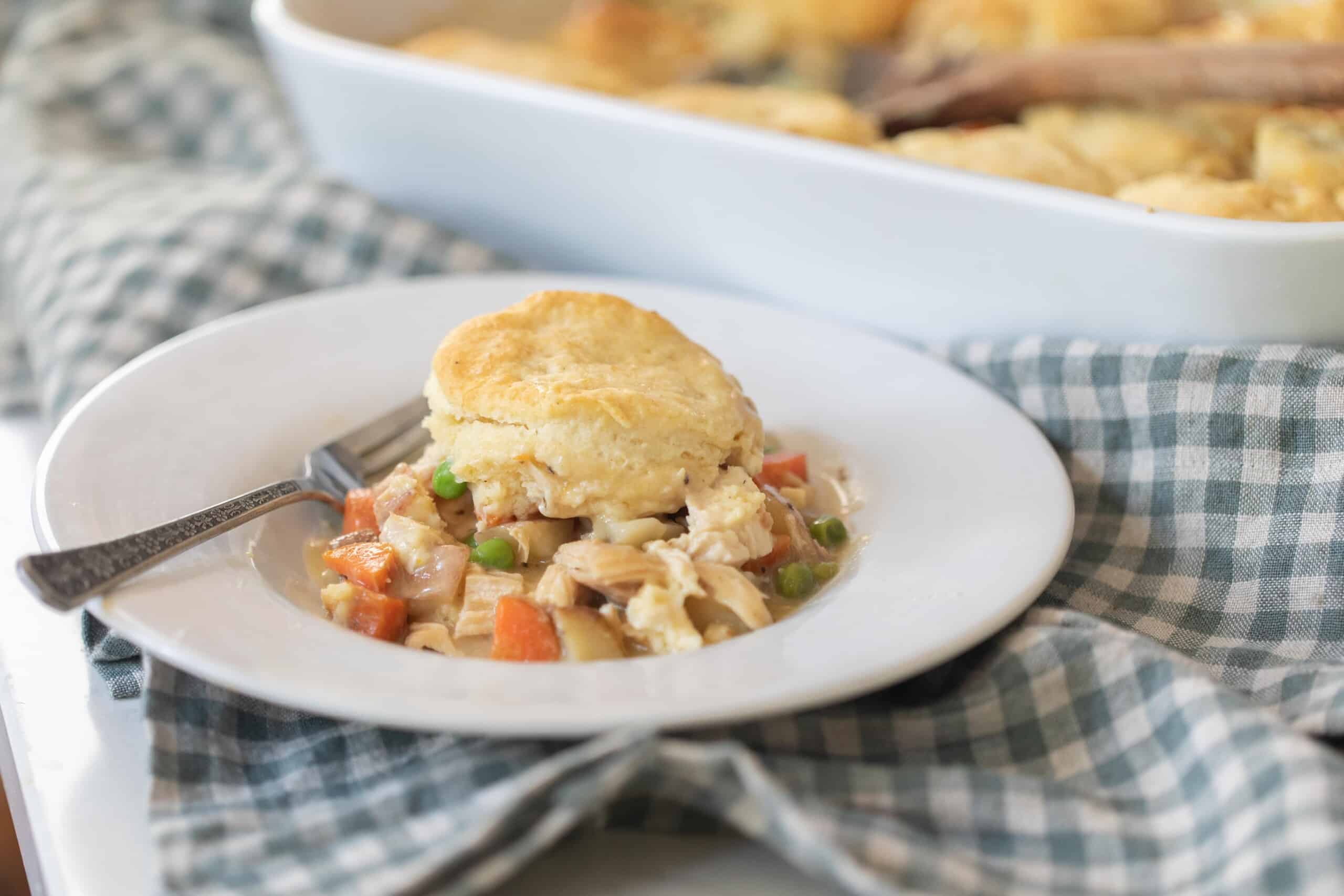 A serving of chicken pot pie on a white plate on a white and blue checked towel with a baking dish or more chicken pot pie in the background