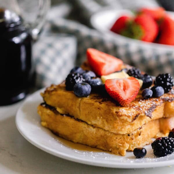 two pieces of sourdough French toast on a white plate and topped with fresh fruit. Maple syrup and strawberries in a bowl are in the background