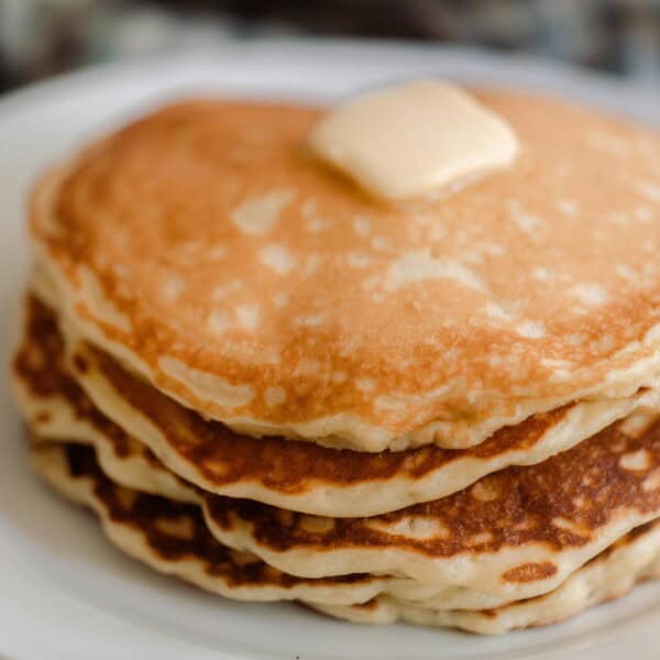 close up picture of sourdough pancakes stacked up 6 pancakes high with a pat of butter and maple syrup. The pancakes are on a white plate