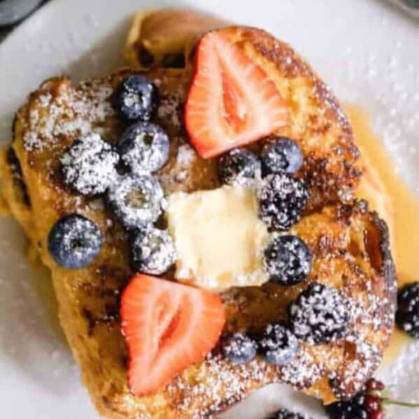 overhead photo of sourdough French toast topped w with blueberries, sliced strawberries, a pat of butter, maple syrup and powdered sugar on a white plate with a bowl of strawberries in the back