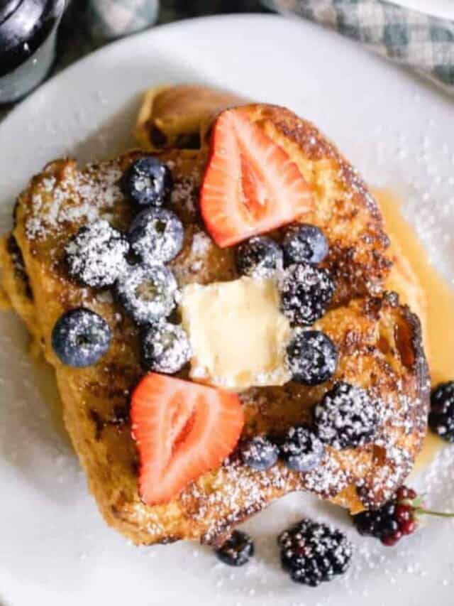 overhead photo of sourdough French toast topped w with blueberries, sliced strawberries, a pat of butter, maple syrup and powdered sugar on a white plate with a bowl of strawberries in the back