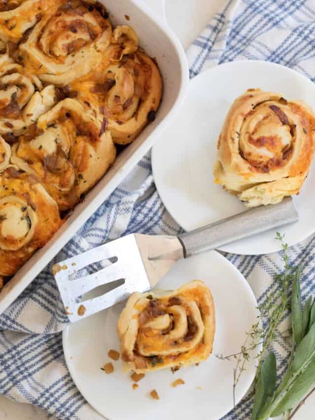 overhead photo of sourdough herb and cheese rolls in a baking dish with two plate with rolls to the right on a top of a blue and white plaid towel