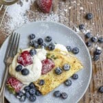 gray plate with one sourdough crepe folded with a bunch of whipped cream topped with blueberries and strawberries. The plate is surrounded by blueberries, strawberries, and a sifter with powdered sugar.