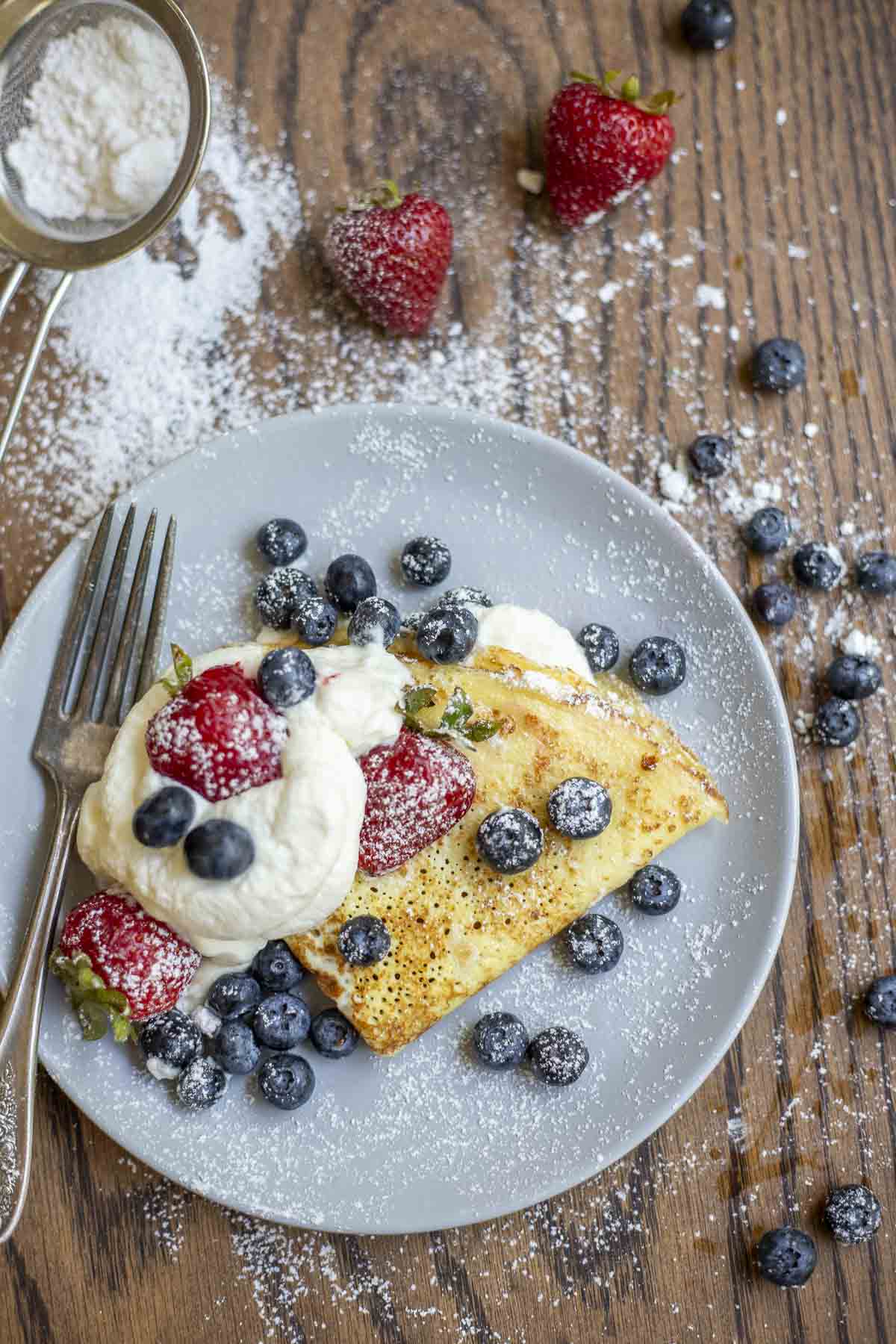 A sourdough crepe on a plate with whipped cream, blueberries, and strawberries. The plate is surrounded by blueberries, strawberries, and a sifter with powdered sugar.