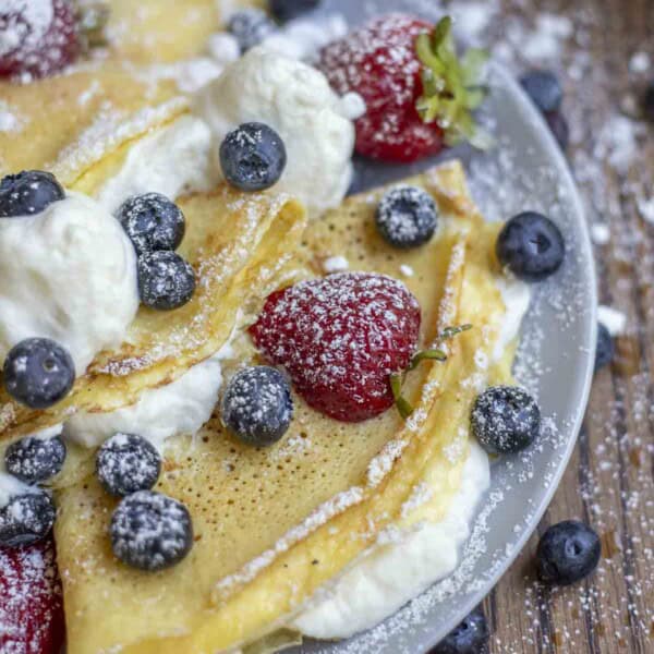 side view of three sourdough crepes with whipped cream filling, blueberries and strawberries on a gray plate on a wood table