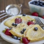 three sourdough crepes topped with blueberries and strawberries on a white plate with a dish of blueberries and a sifter of powder sugar in the background