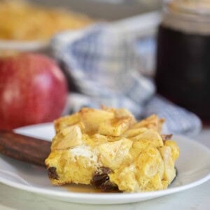 slice of sourdough French toast casserole on a white plate with a wooden spatula. A apple, blue plaid towel and a jar of maple syrup is in the background