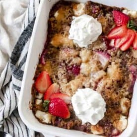 sourdough strawberry cobbler topped with fresh whipped cream and sliced strawberries. The cobbler is in a white baking dish on a white and black stripped towel