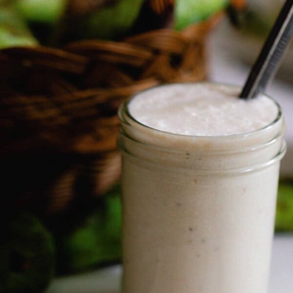 a creamy pawpaw smoothie in a glass mason jar with a metal straw on a white countertop with a basket of pawpaws in the background