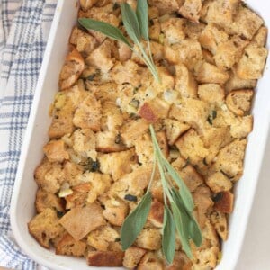 hands holding a white casserole dish full of homemade sourdough stuffing topped with fresh sage