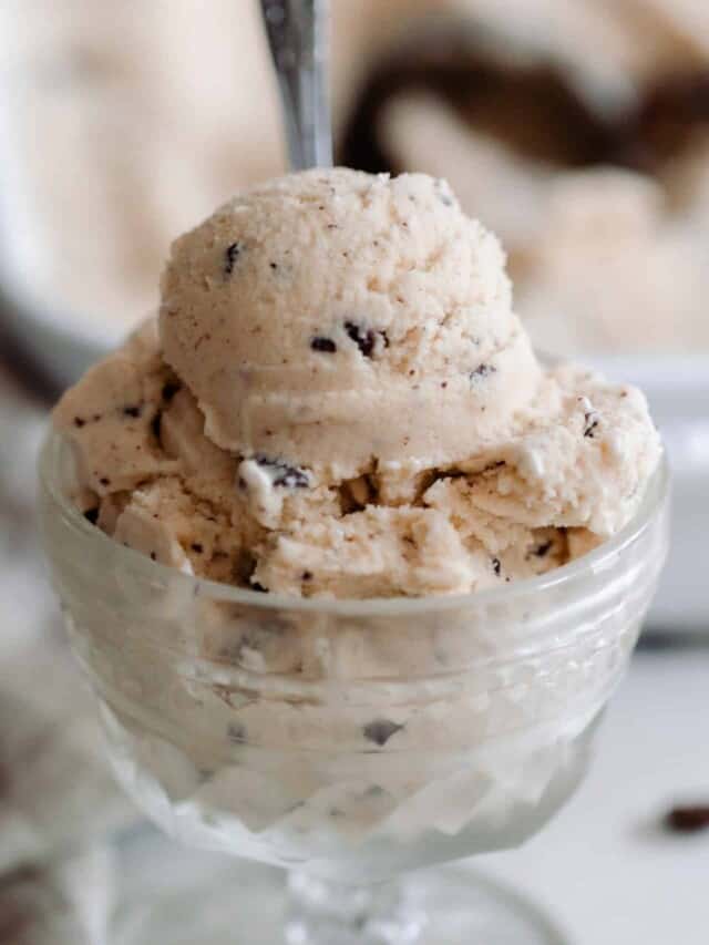 java chip ice cream in a vintage ice cream dish on a white countertop with chocolate chips, a towel and a baking dish of more ice cream in the background