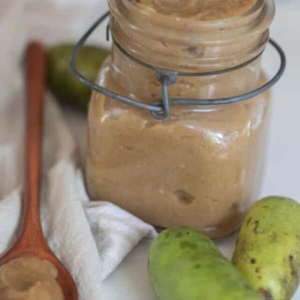 a swing top mason jar with pawpaw jam on a white towel with fresh pawpaw and a wooden spoons with jam in front of the jar