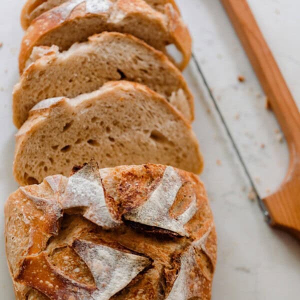 A sourdough boule cut into slices on a white countertop with a bow knife to the side