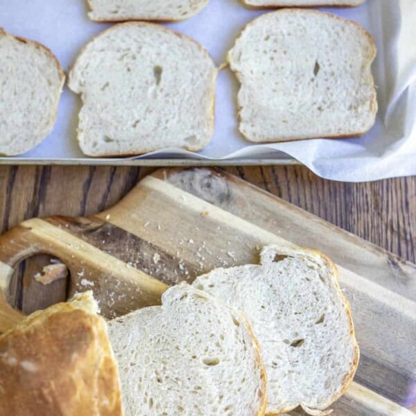 sourdough bread loaf cut on a cutting gbaord with a baking sheet of sliced bread in the background