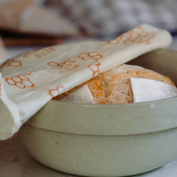 A sourdough loaf in a green bowl with a white and orange bees wax wrap covering it half way