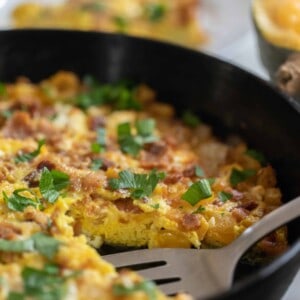 side view of a pumpkin frittata topped with feta, cheddar cheese and fresh herbs in a cast iron skillet with a halved pumpkin in the background