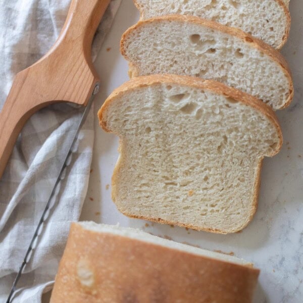 overhead photo of a loaf of sourdough sandwich bread with four slices off on white countertop with a towel and bread knife to the left