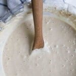 overhead photo of a sourdough starter in a glass bowl with a wooden spoon in the starter. The bowl is next to a white and blue checked towel