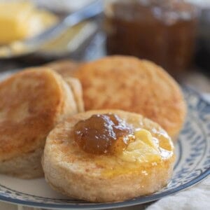 three sourdough crumpets on a blue and white plate topped with butter and jam with more butter and jam in the background