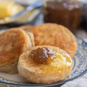 three sourdough crumpets on a blue and white plate topped with butter and jam with more butter and jam in the background