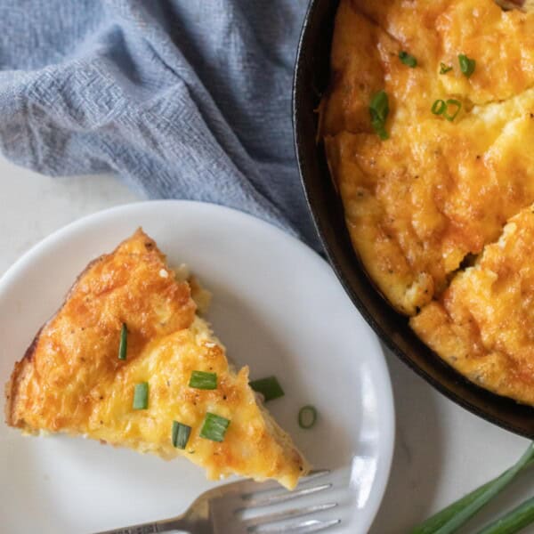 Slice of ham and cheese frittata on a white plate with a silver fork with a cast iron skillet in the background with the rest of the frittata