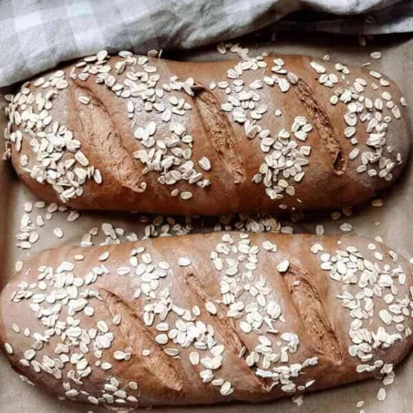 Two loaves of sourdough brown bread dough on parchment paper scored and sprinkled with rolled oats next to a checkered tea towel