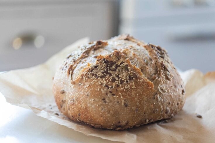 loaf of seeded sroudough bread on parchment paper with a white oven and cabinet int he background