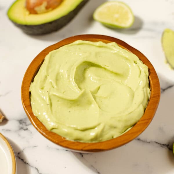 Avocado aioli in a terracotta bowl on a white countertop surrounded by ingredients, such as an open avocado and garlic